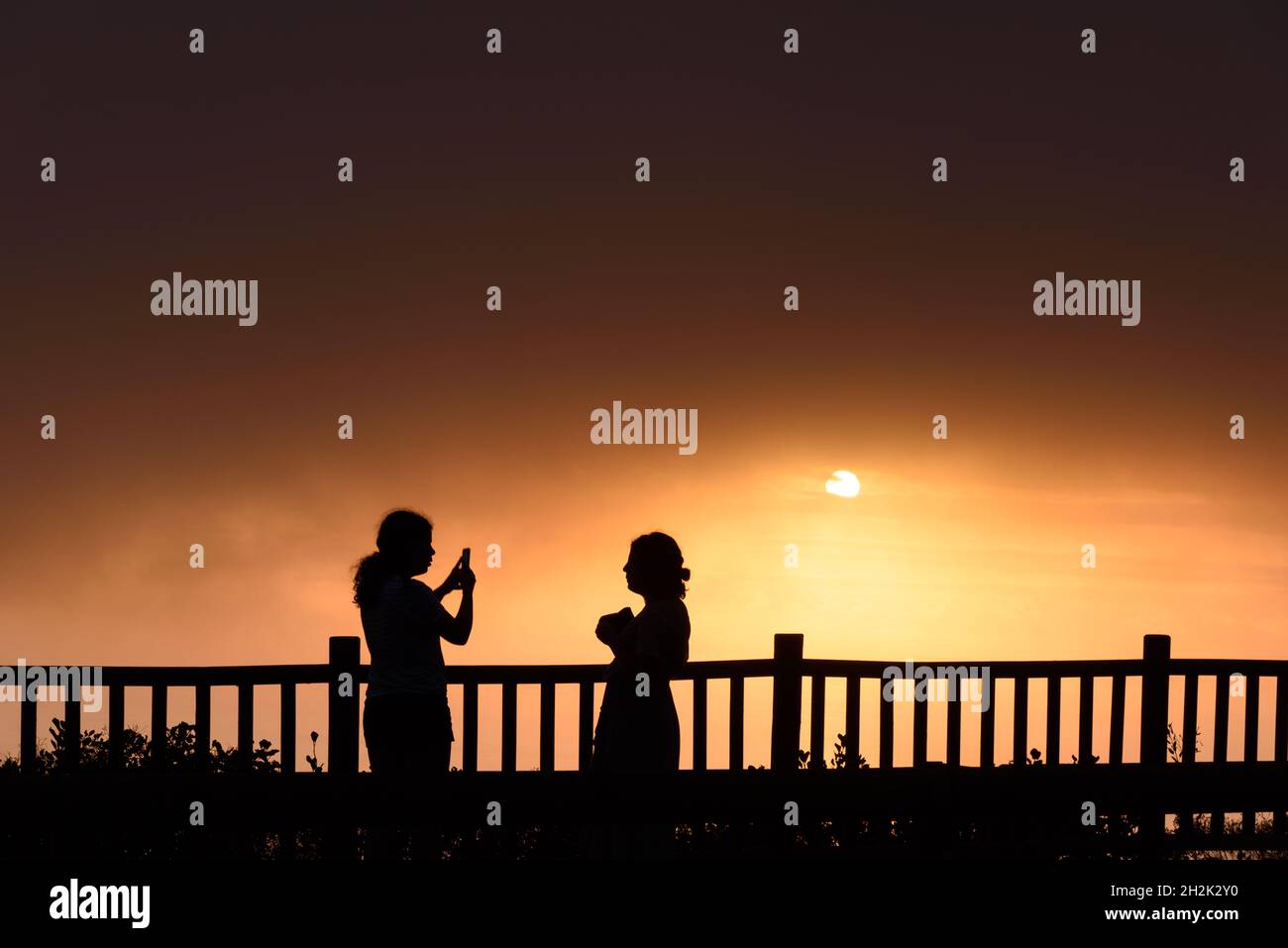 Silhoutte von zwei jungen erwachsenen Frauen, die bei Sonnenuntergang Sonne stehen. Sie machen Fotos Stockfoto