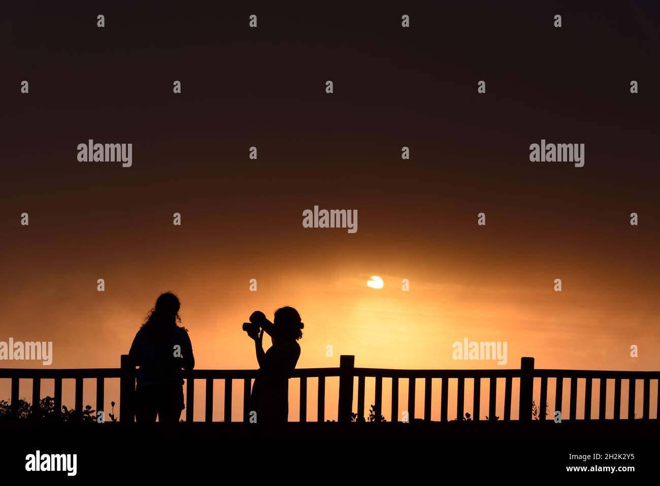 Silhoutte von zwei jungen erwachsenen Frauen, die bei Sonnenuntergang Sonne stehen. Sie machen Fotos Stockfoto