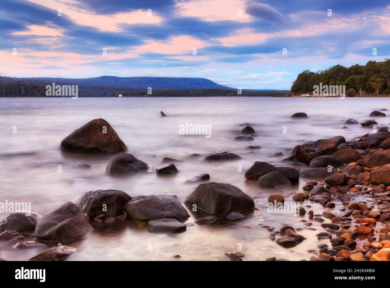 Malerischer Sonnenaufgang über dem verschwommenen Wasser des Lake St Clair mit nassen Uferbrocken und entferntem Fähranleger von der Platypus Bay. Stockfoto