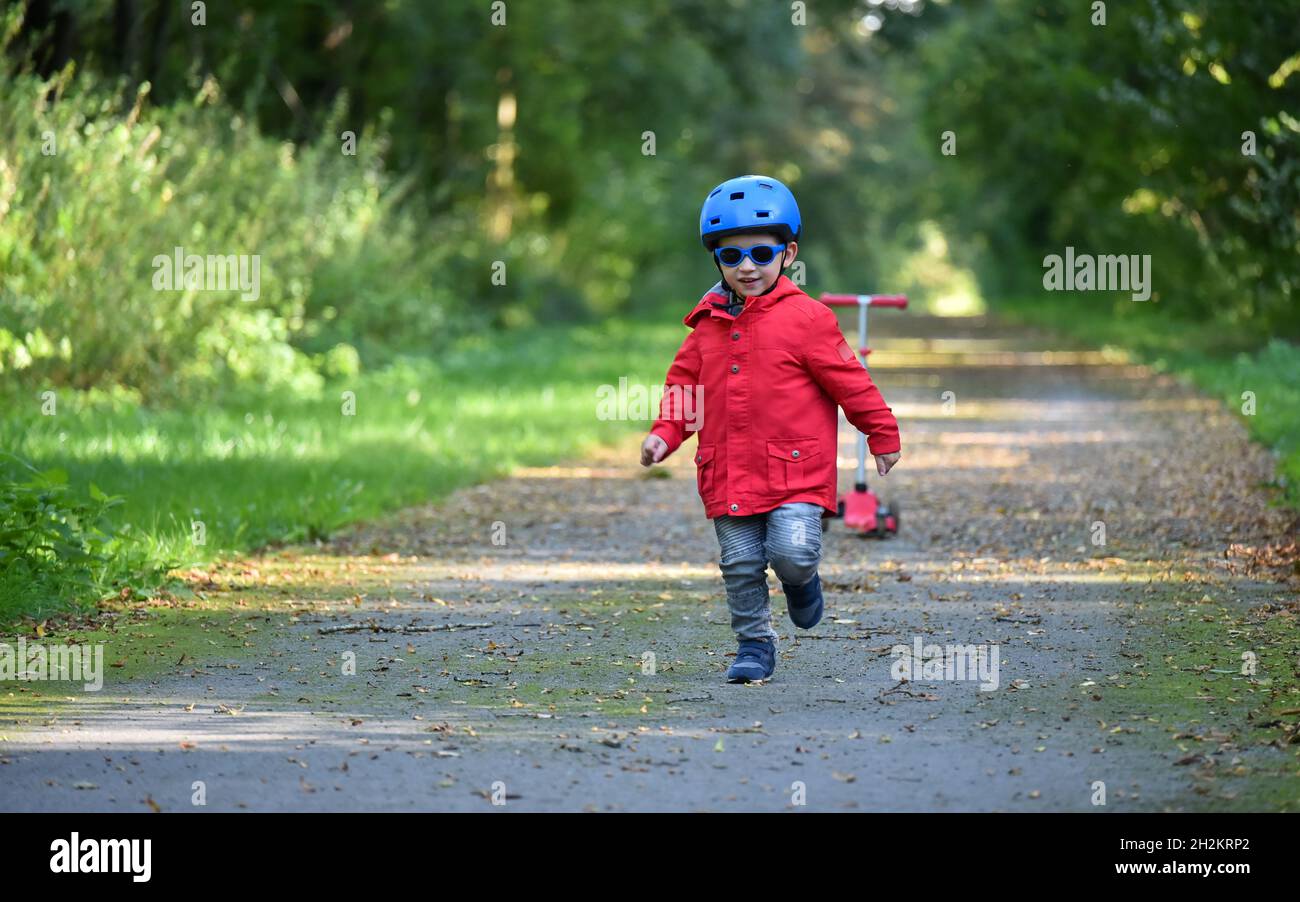 Das kaukasische Kind trägt einen blauen Helm, einen roten Mantel und eine Sonnenbrille und läuft im Herbst auf einem Waldweg. Ein roter Roller ist im Hintergrund; Stockfoto