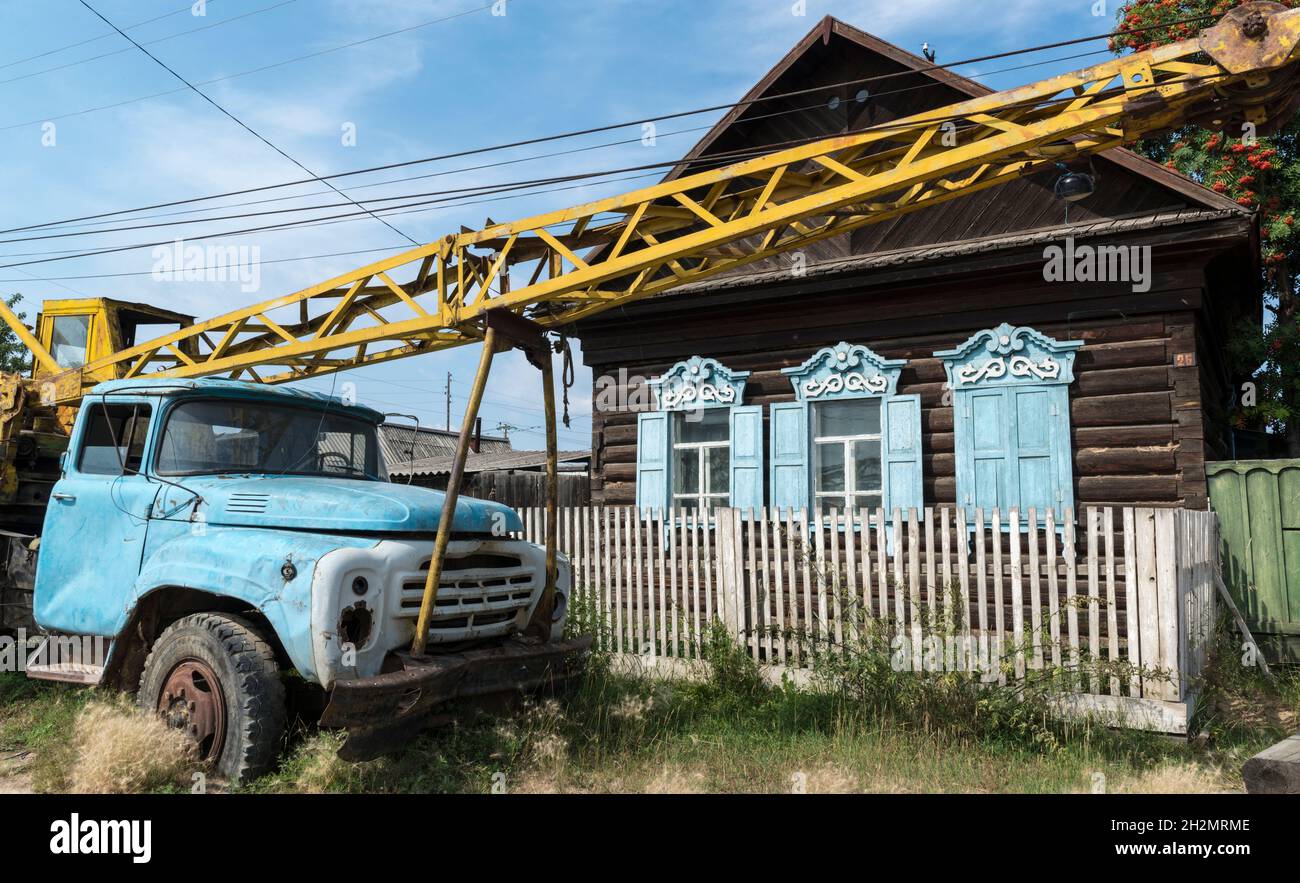 Alte verrostete sowjetische Kranwagen links vor einem traditionellen Holzhaus im Dorf Barguzin. Republik Burjatien, Russland Stockfoto