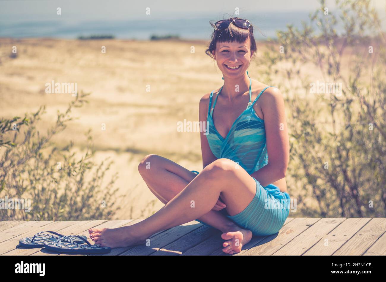 Junge schöne Mädchen in Bikini, pareo wickeln und Sonnenbrille auf dem Kopf sitzen auf Holzweg, lächeln und Blick auf die Kamera, Düne der Litauischen Kurischen Nehrung Stockfoto