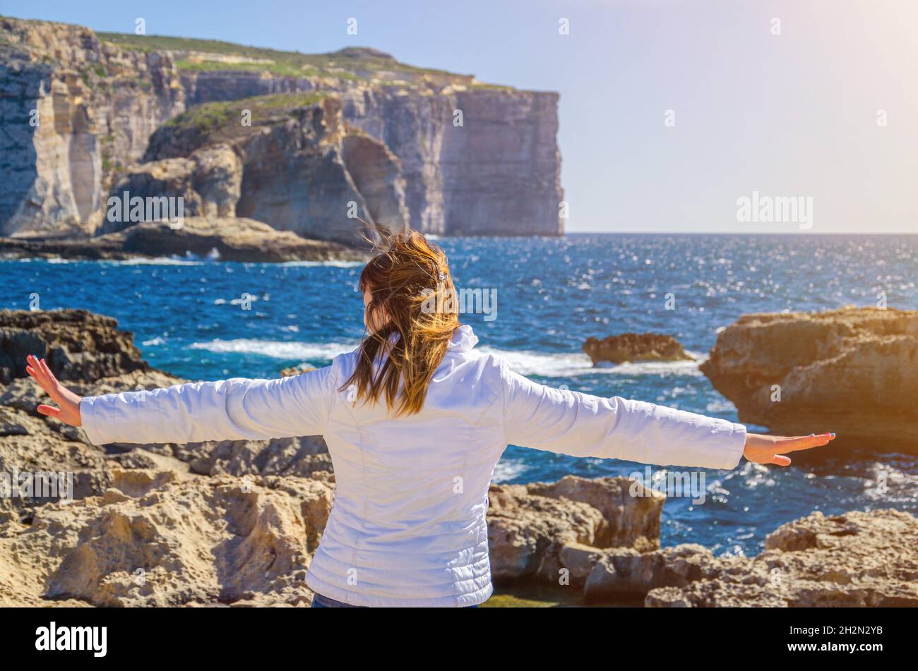 Junge Frau Reisende mit weißer Jacke posiert auf Steinküste der Dwejra Bay, weit heben die Hände und wegschauen Entfernung am Horizont des Mittelmeers se Stockfoto