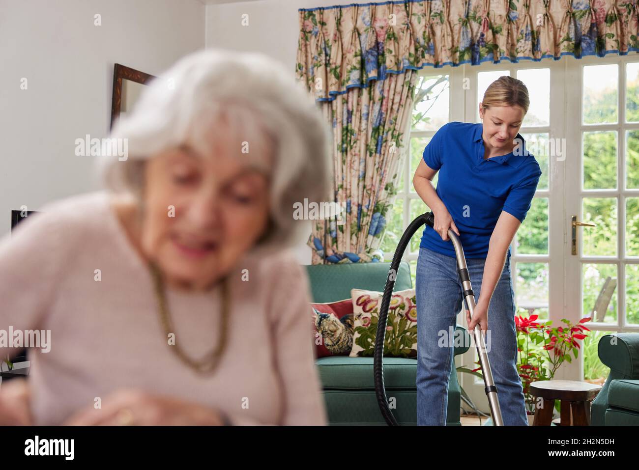 Weibliche Haushilfe Reinigung Haus Mit Staubsauger, Während Ältere Frau Zeitung Liest Stockfoto