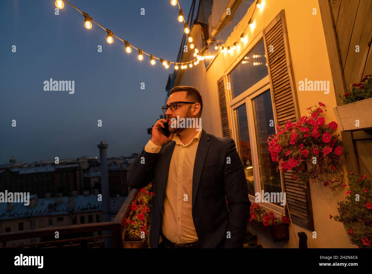 Geschäftsmann, der unter beleuchteten Fadenlampen auf dem Balkon mit einem Smartphone spricht Stockfoto