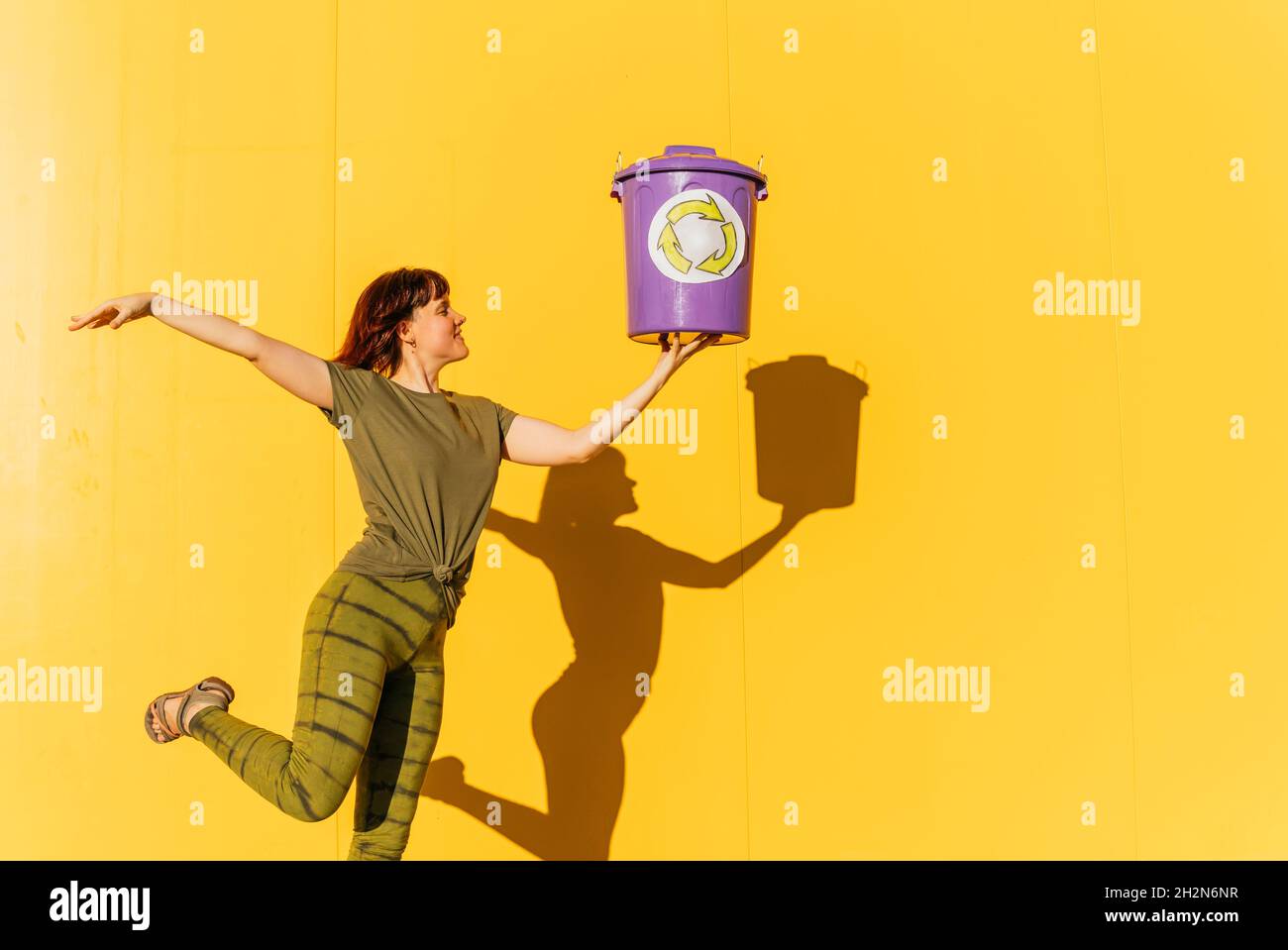Frau mit mittlerem Erwachsenen, die den Abfalleimer in der Hand hält, während sie auf einem Bein vor der gelben Wand steht Stockfoto