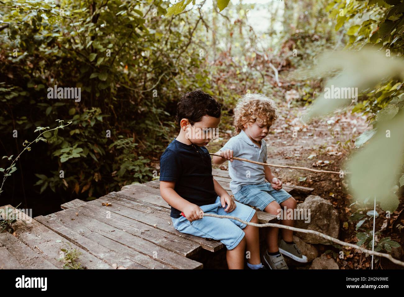 Junge mit einem Freund, der auf einem Steg sitzt, während er im Wald angeln kann Stockfoto
