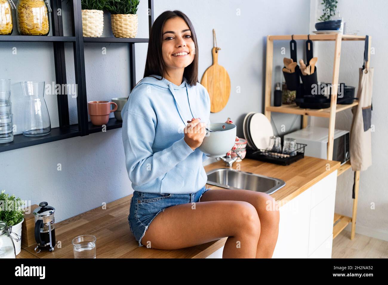 Junge Frau, die zu Hause auf der Küchentheke sitzt und etwas zu essen hat Stockfoto