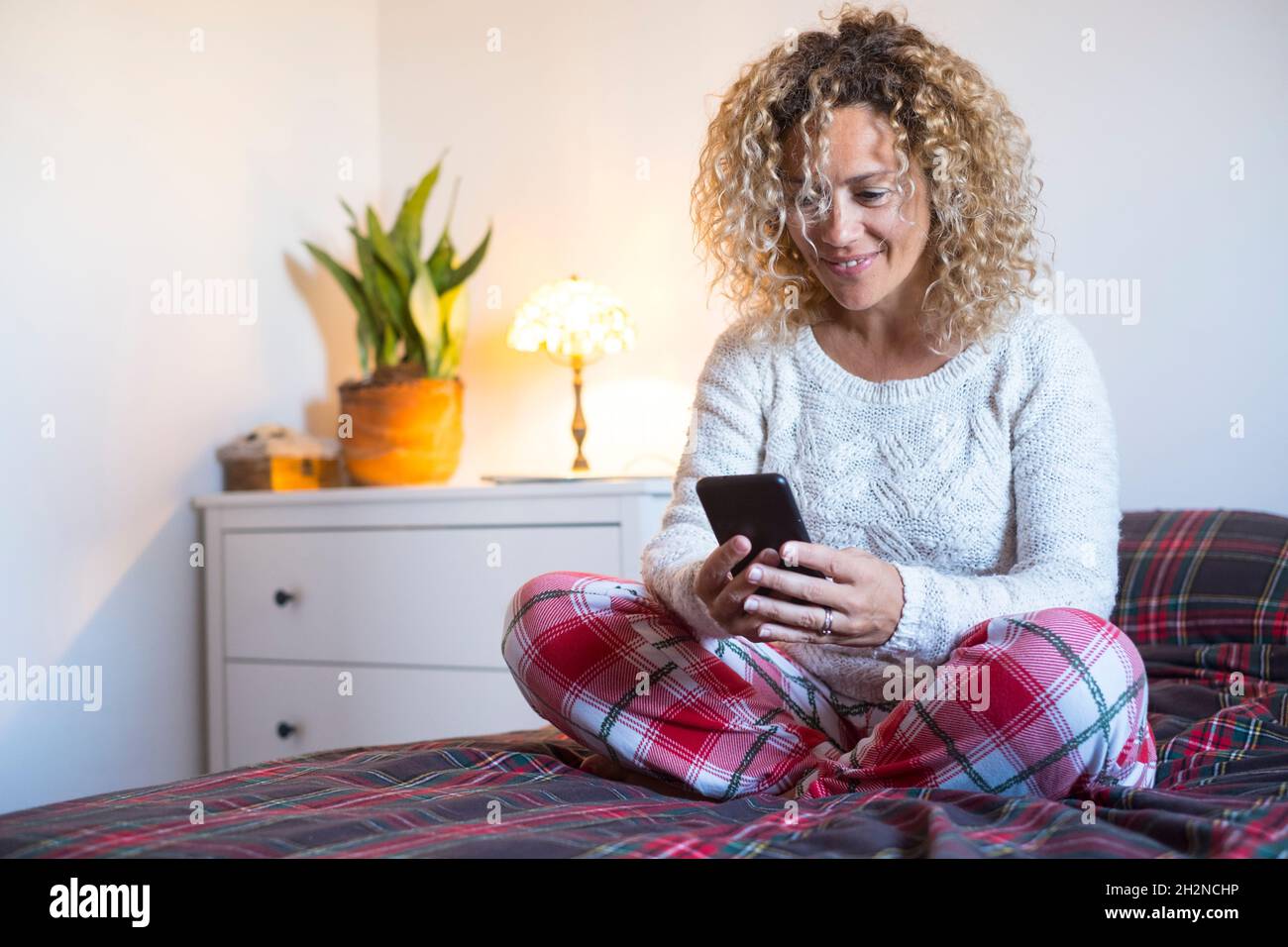 Lächelnde reife Frau, die Smartphone benutzt, während sie mit gekreuzten Beinen auf dem Bett sitzt Stockfoto