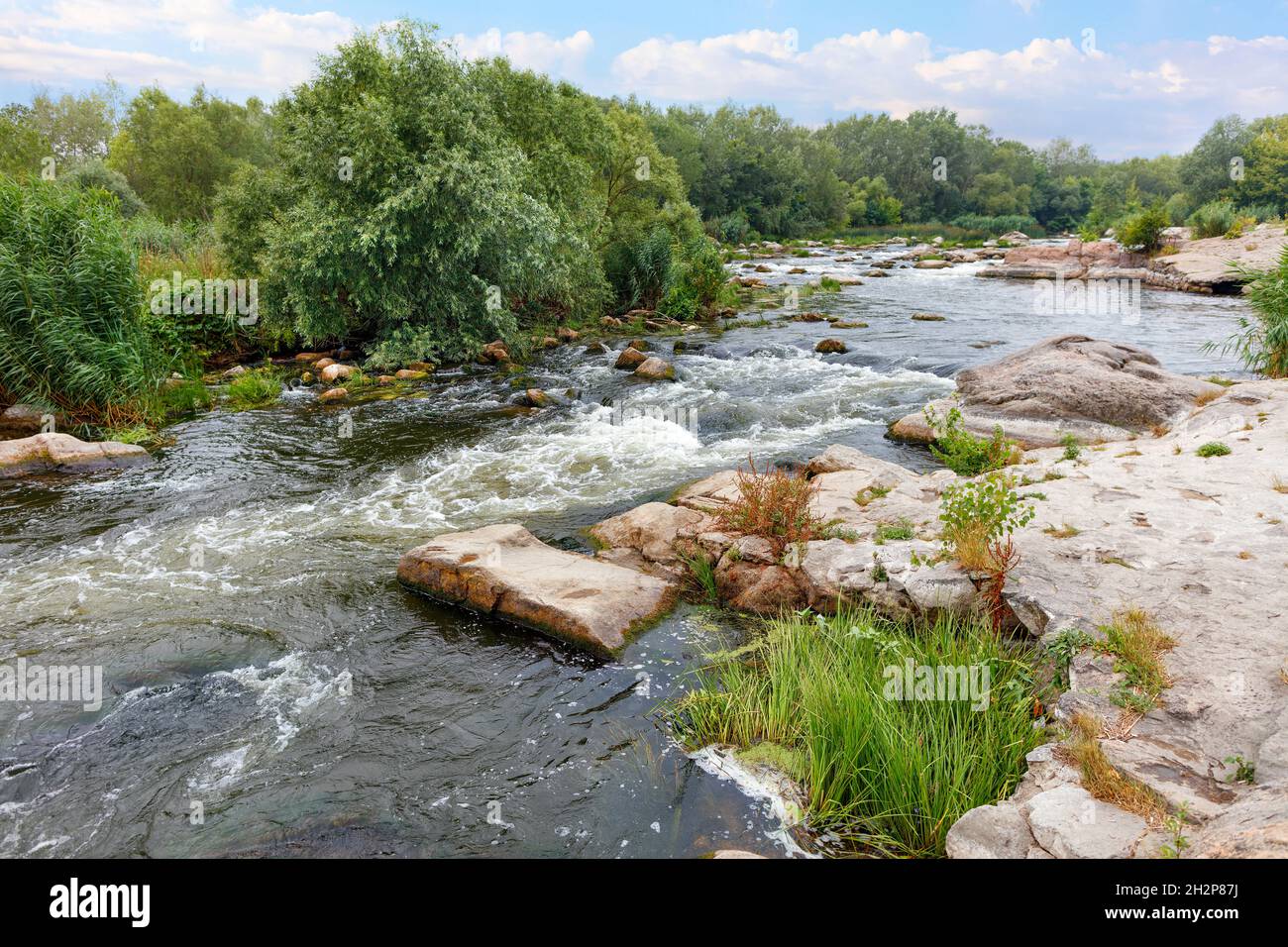 Sommerlandschaft des Flusses zwischen felsigen Küsten, Stromschnellen, Grün und leicht bewölktem blauen Himmel. Stockfoto