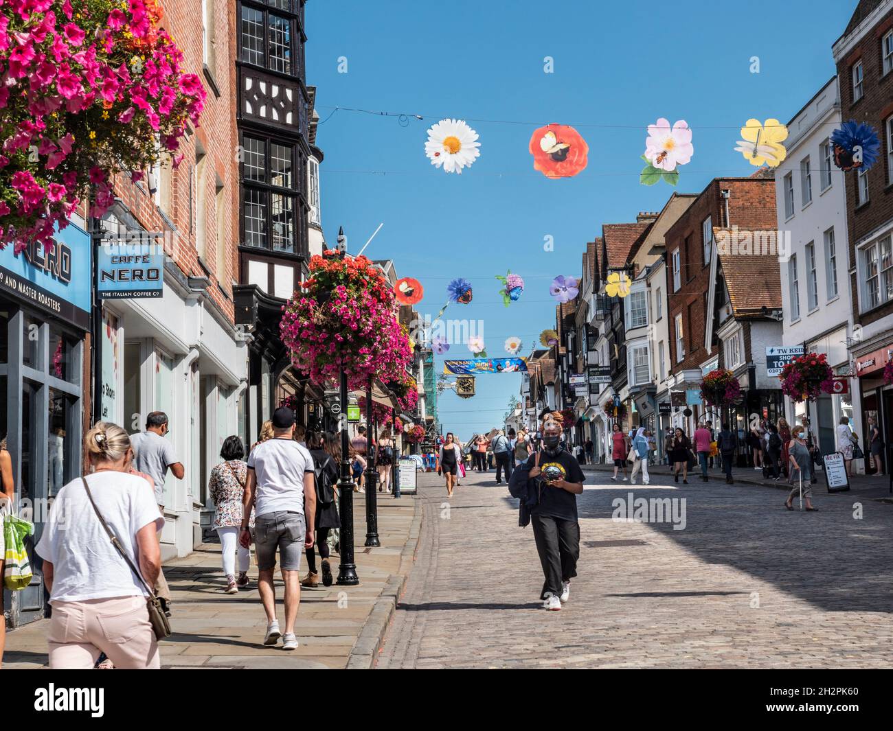 Geschäfte in der Guildford High Street mit Einkäufern, die teilweise Gesichtsmasken tragen. Blumenkörbe im Herbst und farbenfrohe Banner der Saison in klarem, blauem, warmem Sonnenschein Stockfoto