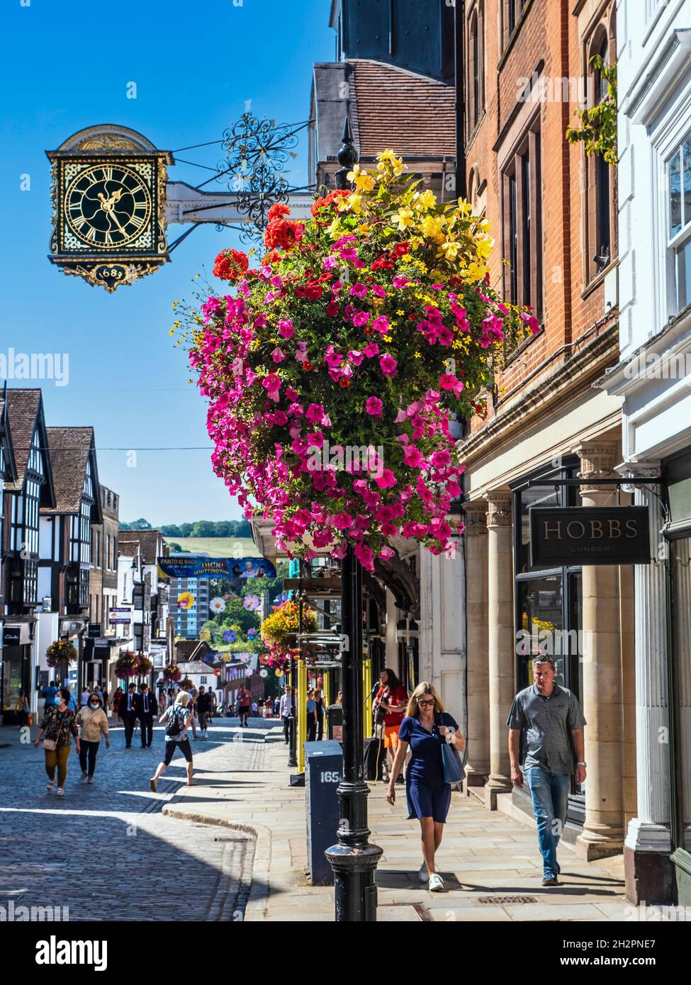 Guildford High Street Geschäfte mit herbstlichen Blumenkörben in klarem blauem warmen Sonnenschein, mit der historischen Guildhall Clock aus dem Jahr 1683 mit frühlingshängenden Blumenkorbblumen im Vordergrund Guildford High Street Surrey UK Stockfoto