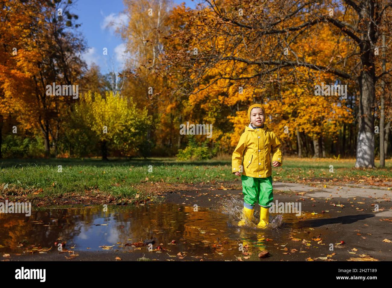 Lächelnder Junge im gelben Regenmantel hat Spaß mit Pfütze Stockfoto