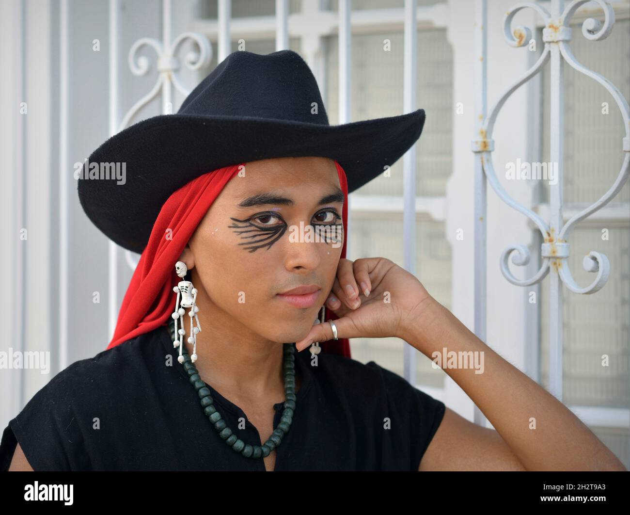 Hübscher junger, nicht binärer Latino mit schwarzem Hut, rotem mexikanischem rebozo und Day of the Dead Skelett-Ohrringen posiert vor dem weißen Fenstergrill. Stockfoto