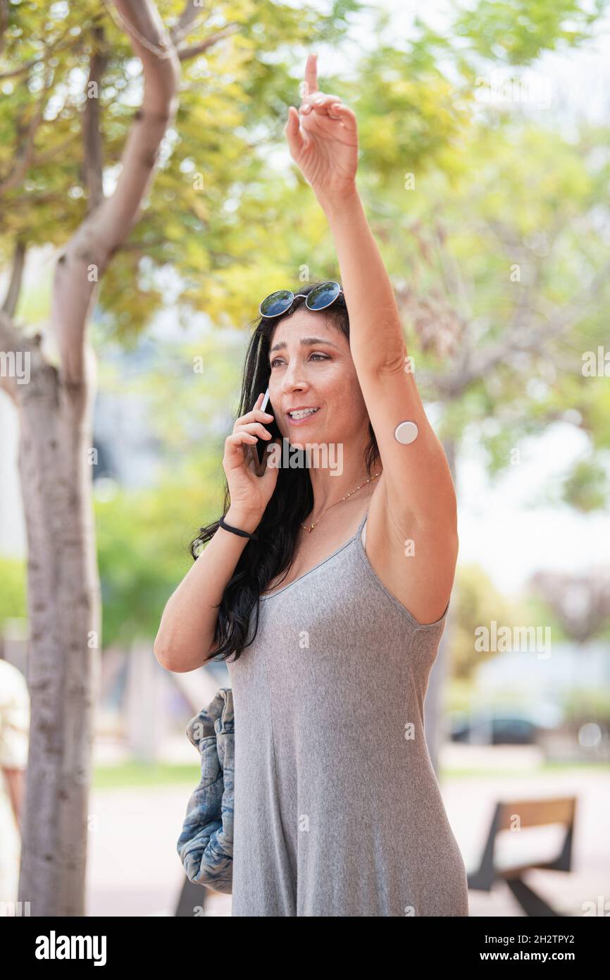 Erwachsene Frau mit einem Glukosesensorgerät auf ihrem linken Arm, die an einem sonnigen Tag telefoniert und ein Taxi ruft Stockfoto