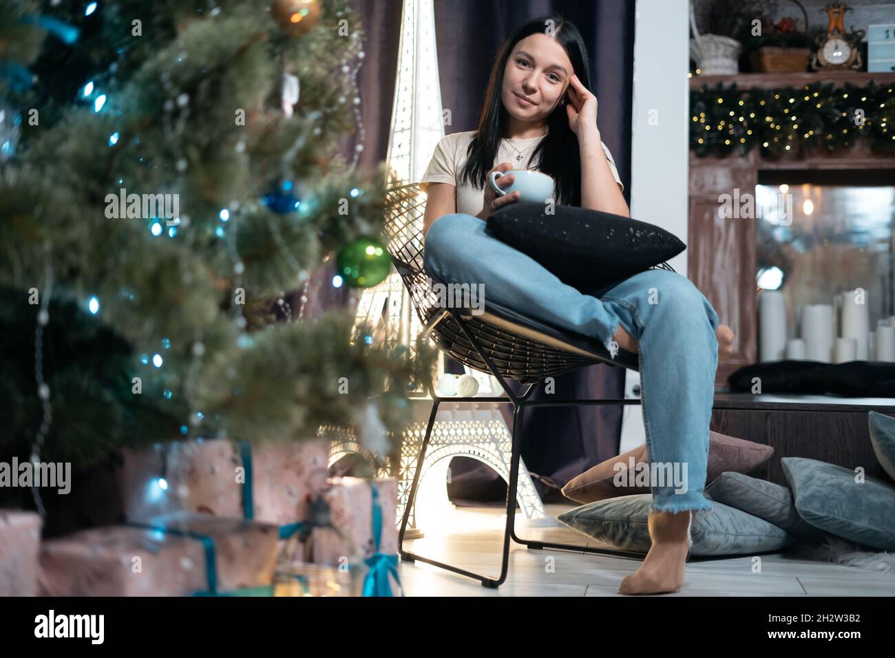 Junge Frau trinkt Kaffee und ruht sich zu Hause mit einer Tasse am Weihnachtsbaum aus Stockfoto