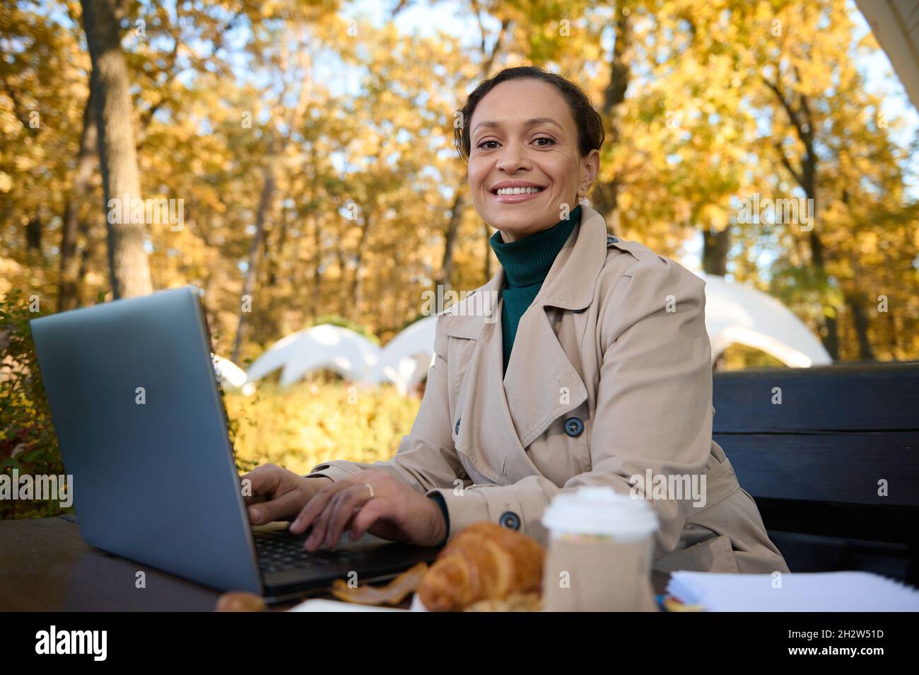 Attraktive Frau mit einem schönen Lächeln, Geschäftsfrau, Freiberufler arbeitet am Laptop und genießt die Außenarbeit im Eichenhain mit Bastelpapier Stockfoto