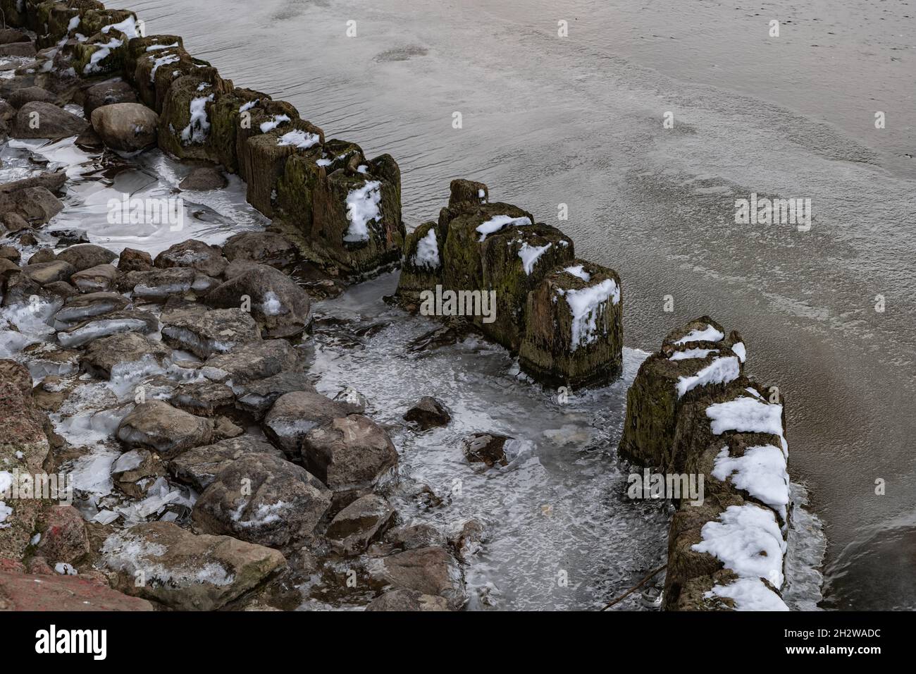 Gefrorener Fluss mit alten Holzpfosten an felsiger Küste, kalte Wintersaison. Stockfoto