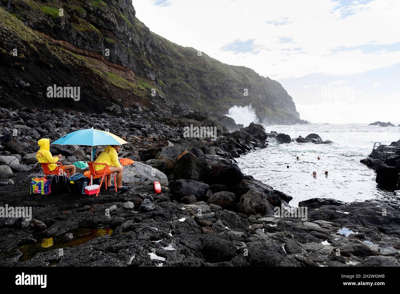 Rettungsschwimmer, die unter einem Regenschirm sitzen und Badenden in vulkanischen Lavaformationen an der Küste von Ferraria, Sao Miguel, Azoren, Portugal, zusehen Stockfoto