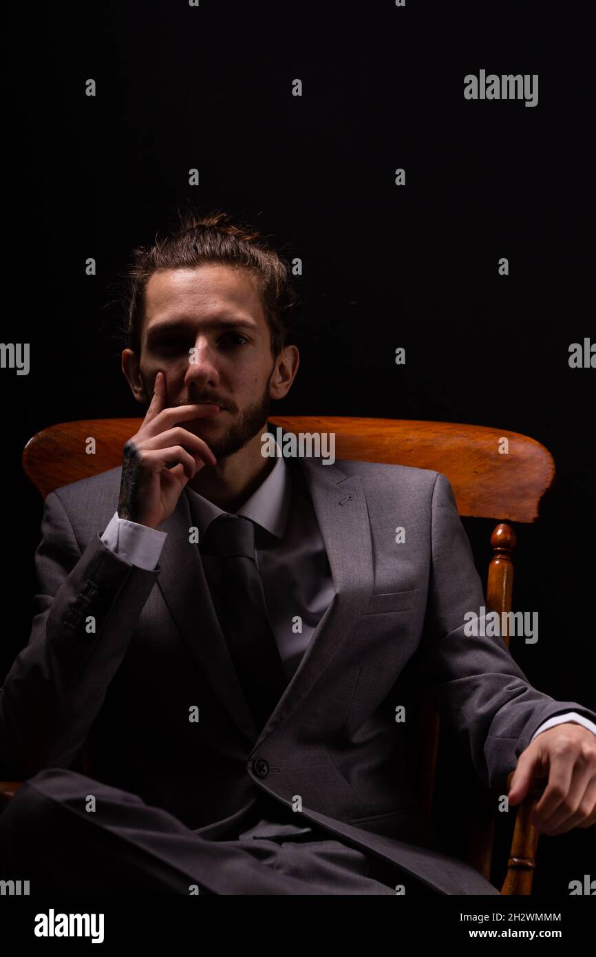 Ein junger Geschäftsmann in einem eleganten grauen Anzug sitzt in einem hölzernen Sessel mit stimmungsvoller und stimmungsvoller Beleuchtung Stockfoto