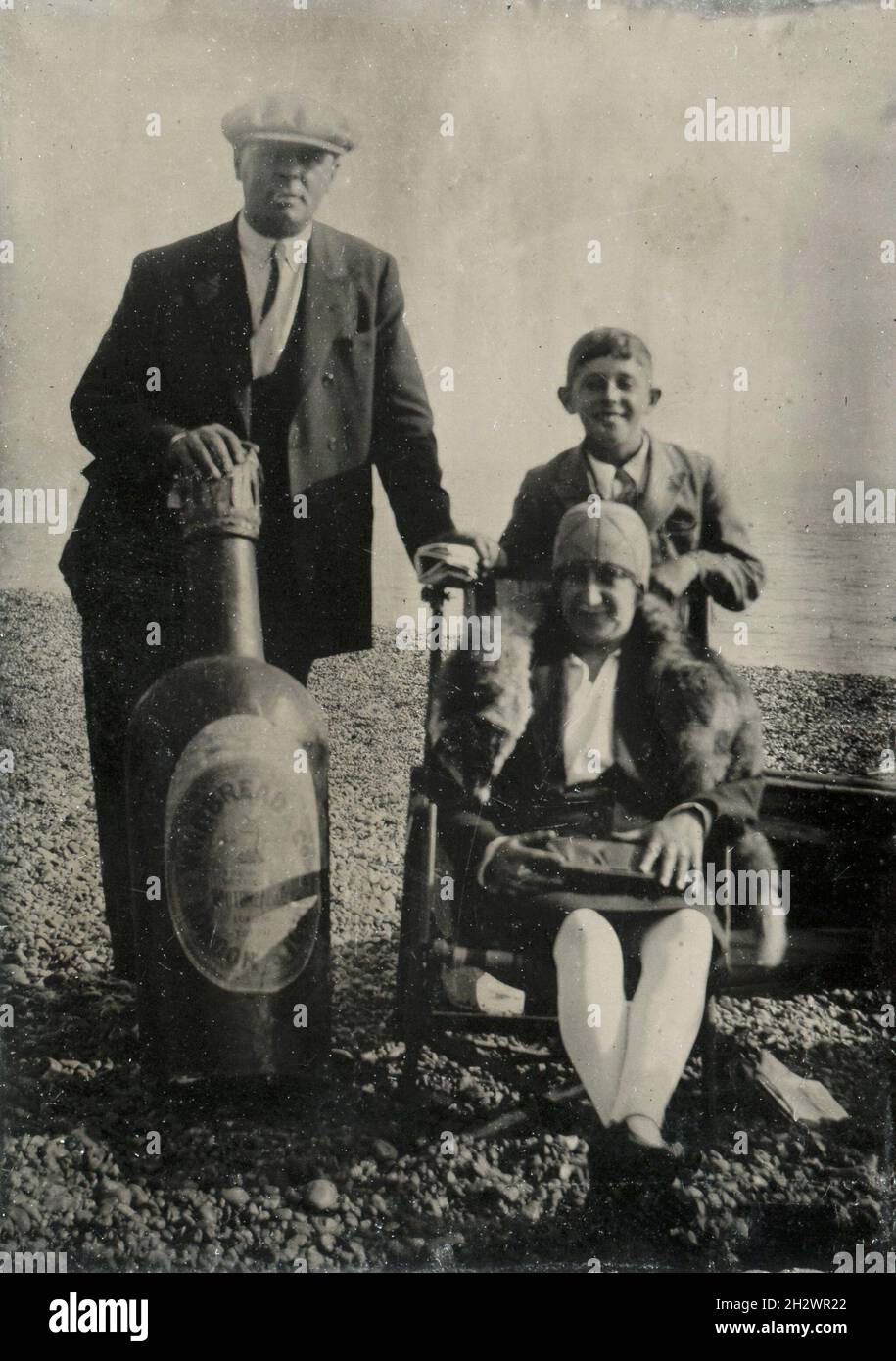 Ein amüsantes englisches „Tintype“-Foto ‘den späten 1920er Jahren, das eine Familie am Strand zeigt. Die Mutter trägt eine Pelzsohle und sitzt auf einem Liegestuhl mit ihrem Sohn hinter ihr. Der Vater hält eine riesige Bierflaschenstütze von ‘Whitbread & Co. London Stout’. Stockfoto