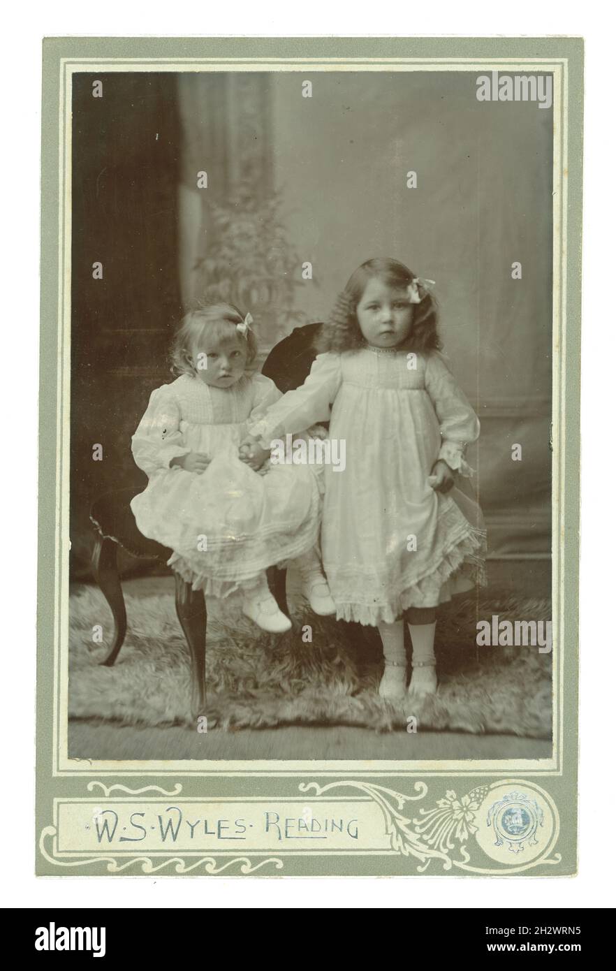 Edwardianische Kabinettkarte Studioportrait von 2 attraktiven entzückenden jungen Mittelklasse-Kindern, Mädchen, die weiße Kleider tragen und die Hände zusammenhalten. Das älteste Mädchen hat ihre Haare gekräuselt und beide tragen Bänder im Haar. Aus dem Fotostudio von W.S. Wyles, Reading, Großbritannien um 1905. Stockfoto
