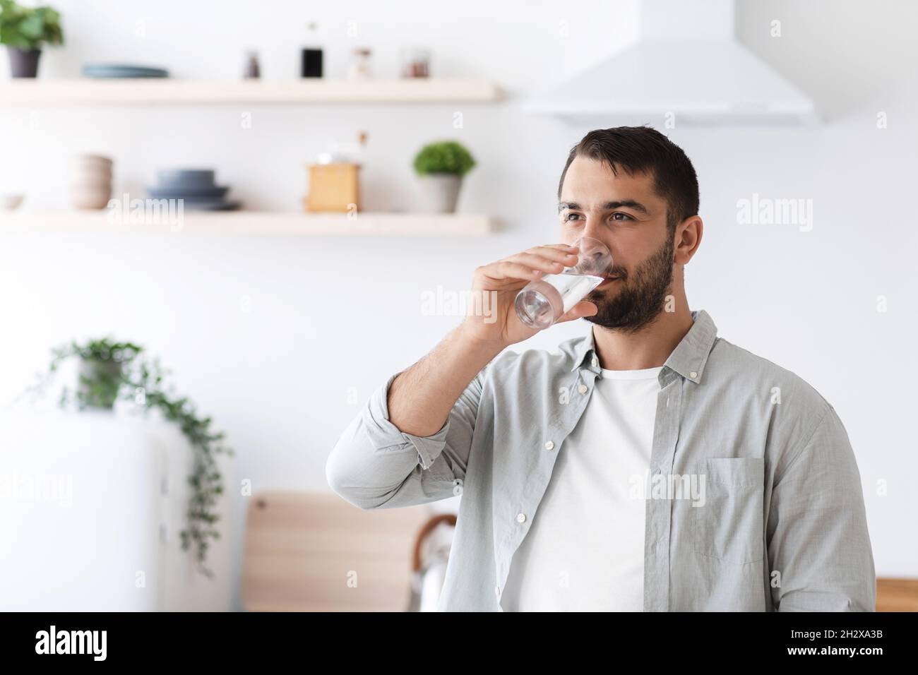 Attraktiver europäischer Mann mittleren Alters in legerem Trinkwasser aus Glas auf minimalistischem Kücheninterieur Stockfoto