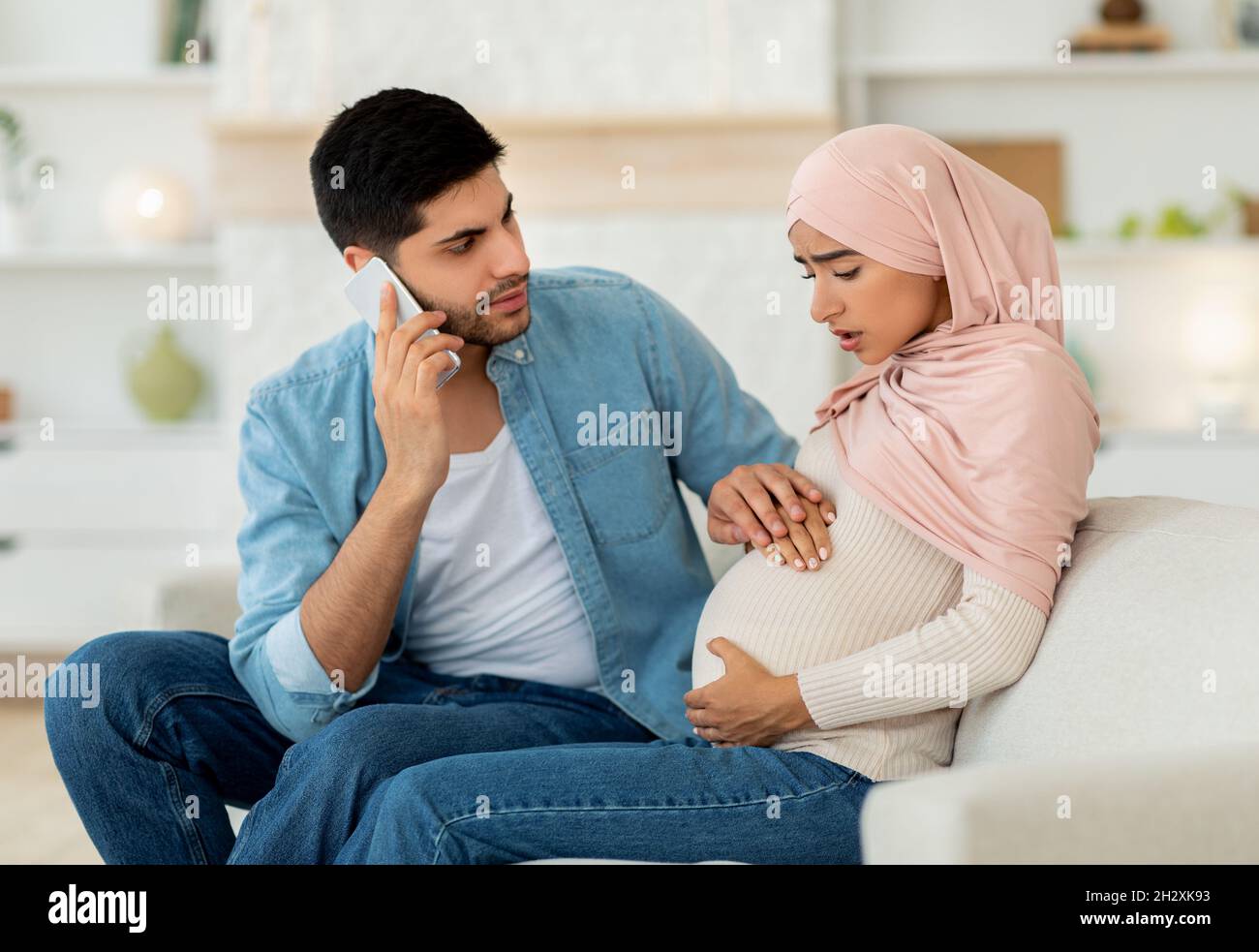 Schwanger arabische Frau mit pränatalen Kontraktionen, besorgt Ehemann rufen Arzt, Handy, häusliche Einrichtung Stockfoto