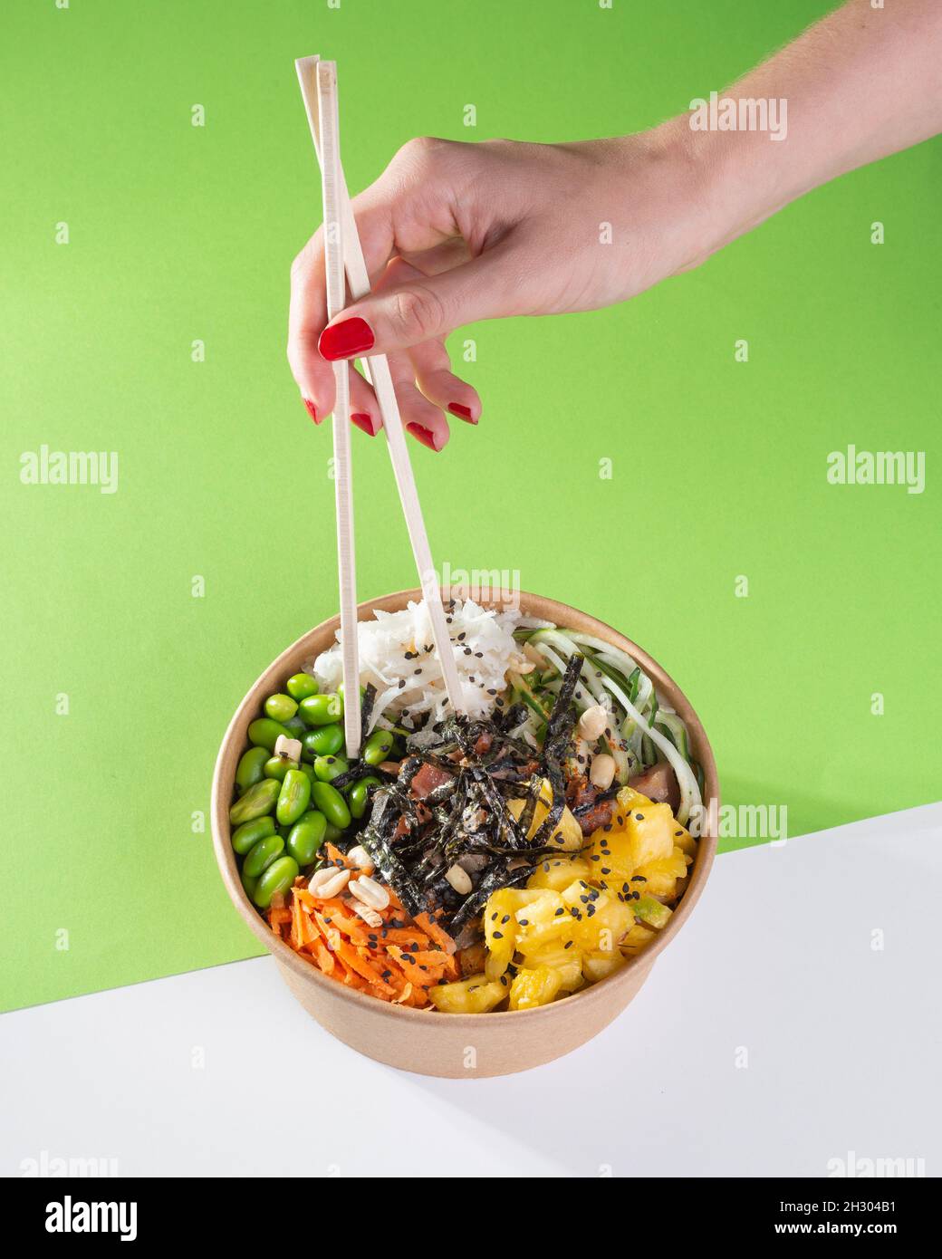 Eine Frauenhand mit Essstäbchen über einer Salatschüssel und Thunfisch, Karotte, Gurke und Ananas mit grünem Hintergrund. Weißer Boden Stockfoto