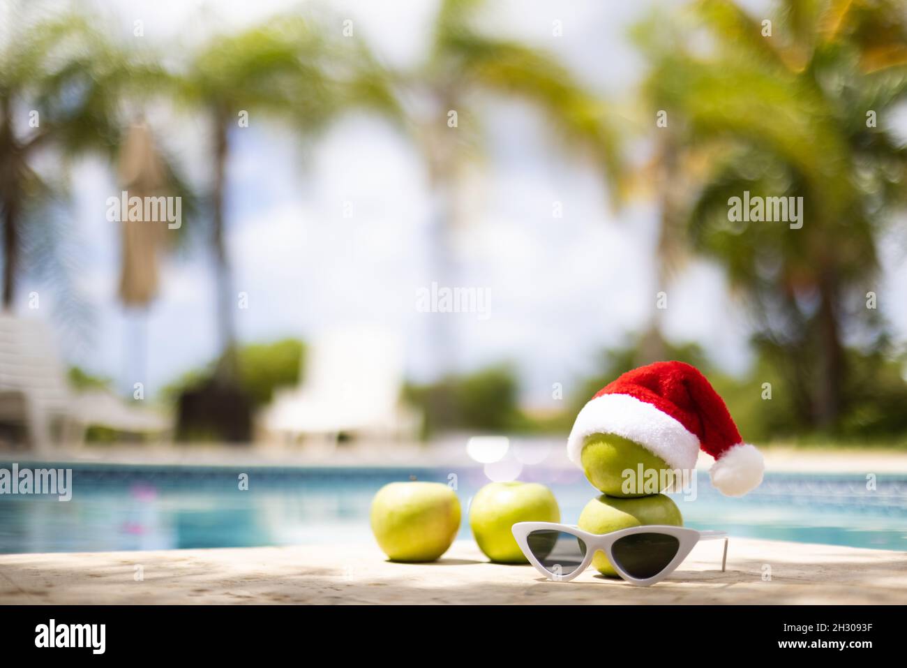 Schneemann aus Äpfeln im Weihnachtshut, der in der Nähe des Schwimmbades mit Sonnenbrillen auf dem Palmenhintergrund steht. Selektiver Fokus, unscharfer Hintergrund. Stockfoto