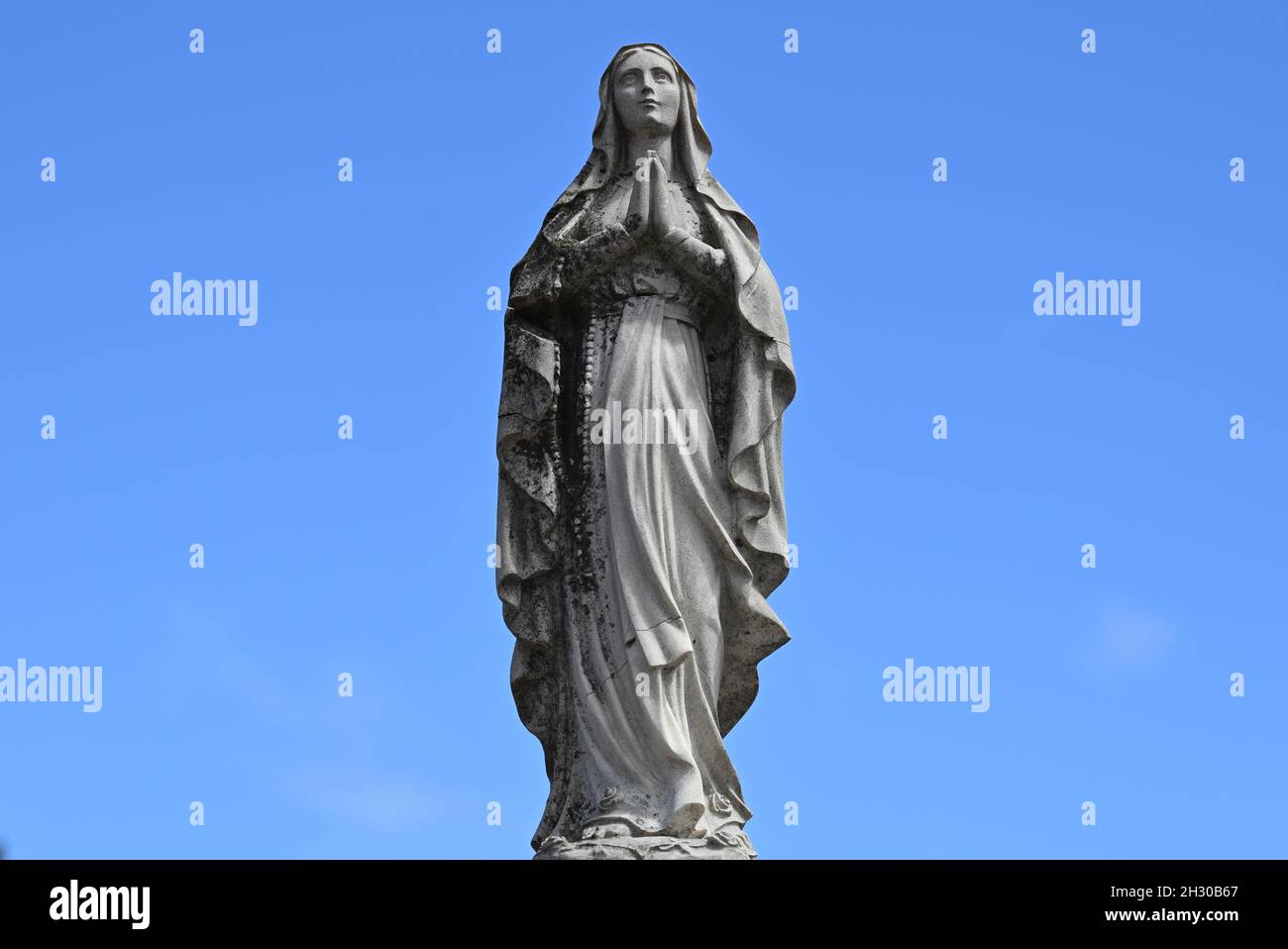 Abgenutzte und rissige Steinskulptur einer verschleierten Frau, die während des Gebets einen Rosenkranz in der Hand hält, mit einem strahlend blauen Himmel im Hintergrund Stockfoto