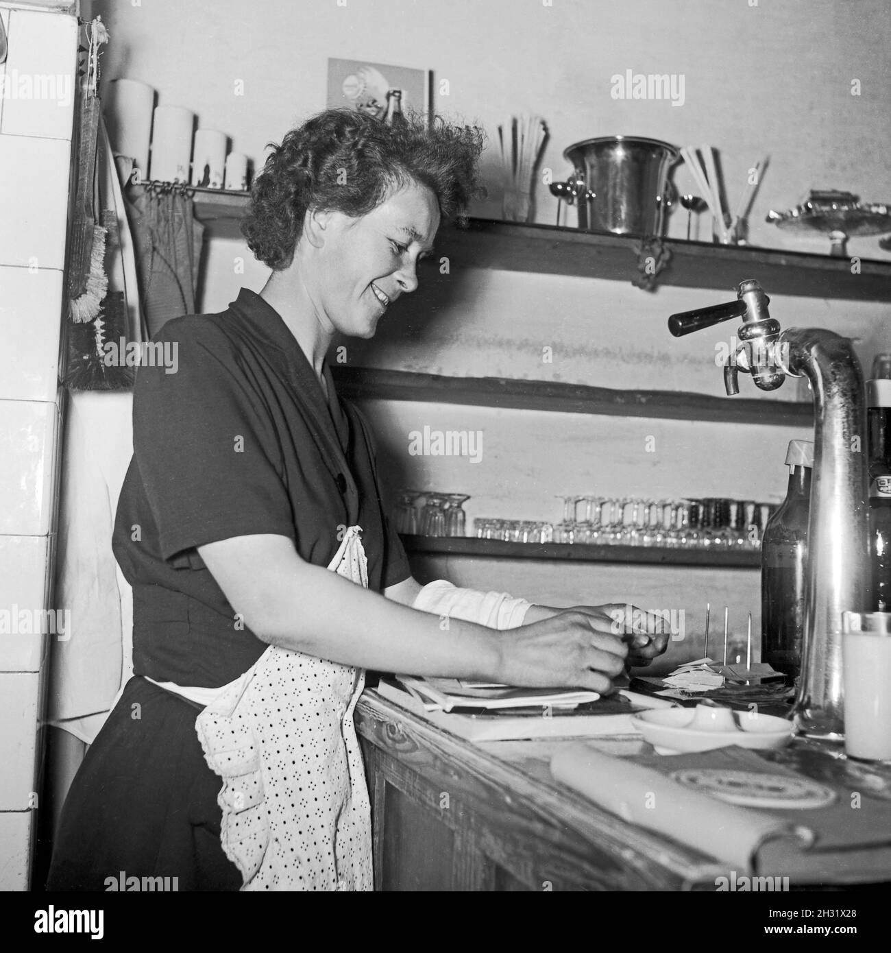 Eine Bedienung ordnet die Bestellungen an der Theke im Gasthaus, Deutschland 1955. Eine Kellnerin, die die Bestellungen am Schalter eines Gasthauses überprüft, Deutschland 1955. Stockfoto