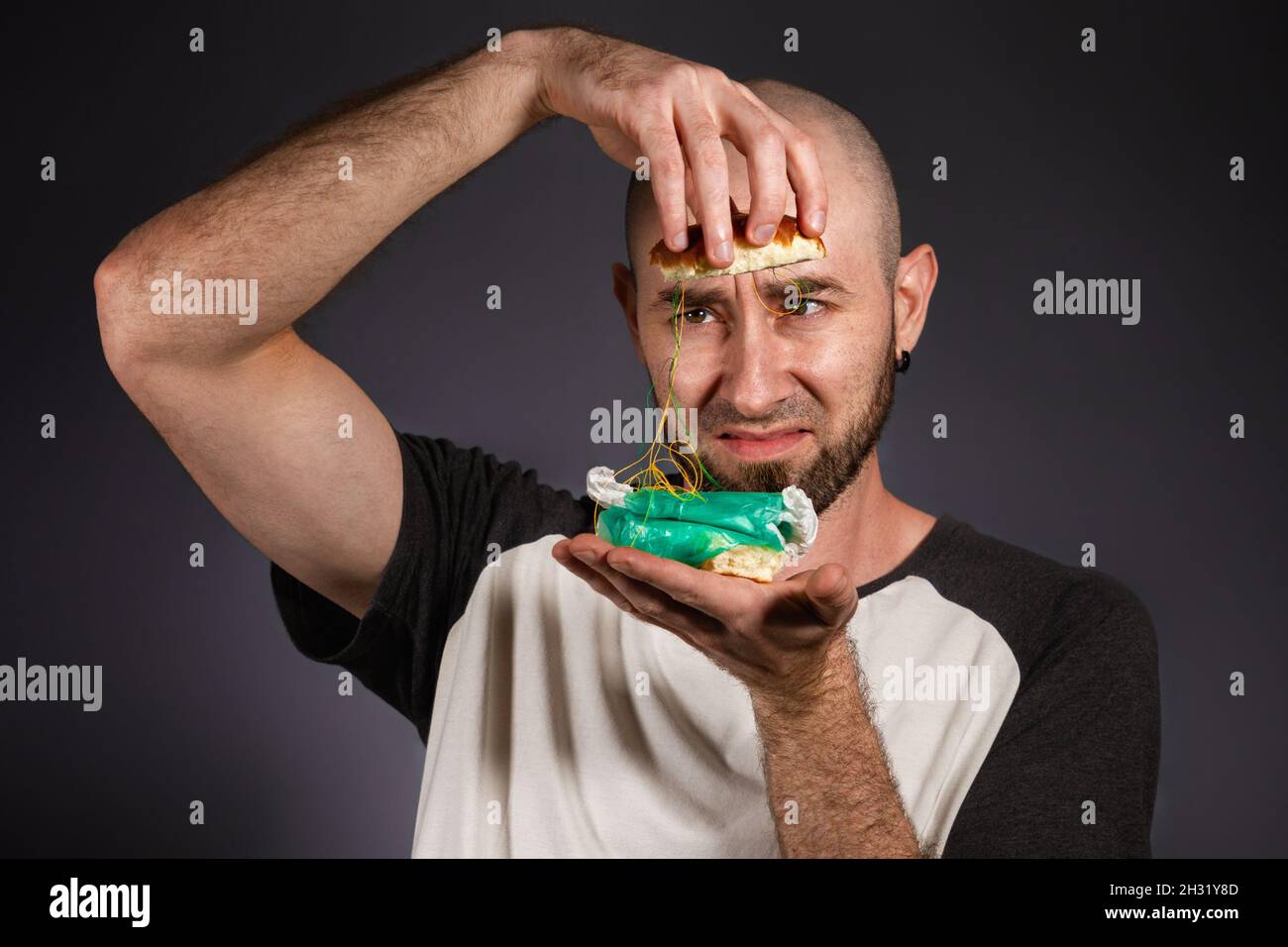 Das Konzept von Fast Food und Ökologie. Ein kahlköpfiger Mann mit Bart blickt entsetzt auf die Füllung eines zerlegten Hamburgers. Dunkler Hintergrund. Stockfoto