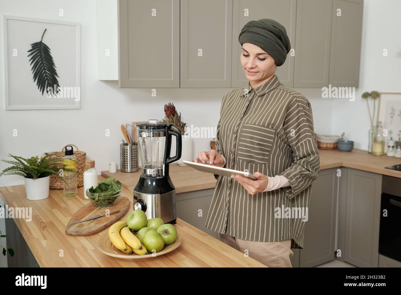 Moderne junge Frau mit Tablet, die online Rezepte von Smoothie durchschaut, während sie am Küchentisch steht Stockfoto