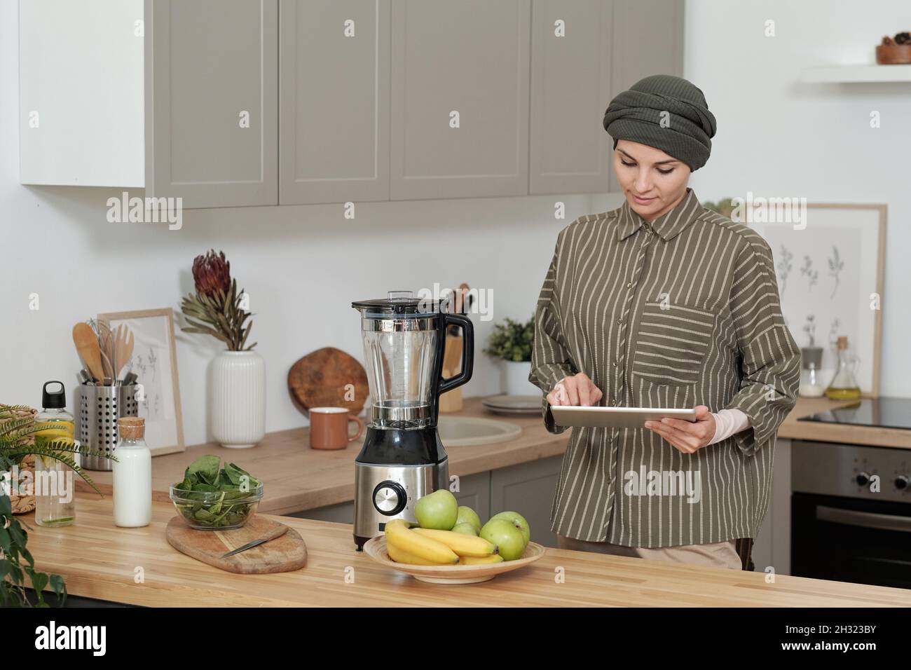 Junge Frau in Casualwear, die Online-Rezepte durchschaut, während sie am Küchentisch steht Stockfoto