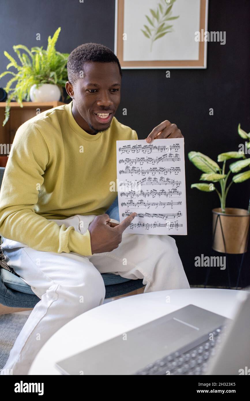 Fröhlicher junger Musiklehrer, der auf Papier mit Notizen zeigt, während er den Schülern während des Online-Unterrichts die Themen erklärt Stockfoto