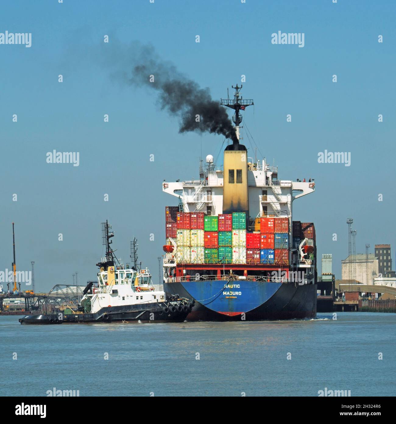 Ankommender Frachtcontainer Frachtschiff & schwarzer Rauch gestapelt mit Ladung Transportcontainer Schlepper schiebt in Richtung Kräne Felixstowe Port Suffolk UK Stockfoto