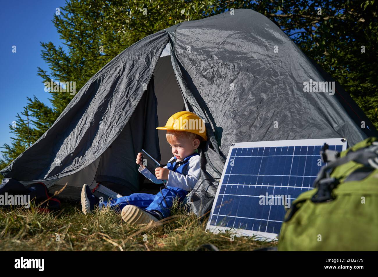 Junger Militärangehöriger 3 Jahre alt, der sich in der Nähe des Zeltes im Lager ausruhte und tagsüber Handytechnologie zum Spielen verwendete, Junge in Uniform und Helm, der auf der Website nach Online-Cartoons schaute Stockfoto