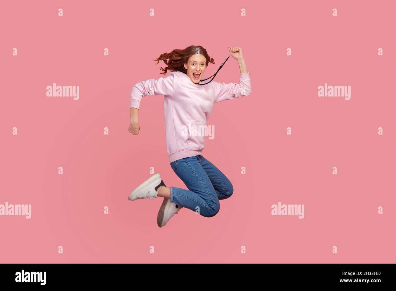 In voller Länge Porträt von extrem glücklich Teenager-Mädchen mit lockigem Haar im Hoodie Springen vor Freude oder Fliegen, feiern Erfolg, Sieg, Indoor-Studio Schuss isoliert auf rosa Hintergrund Stockfoto