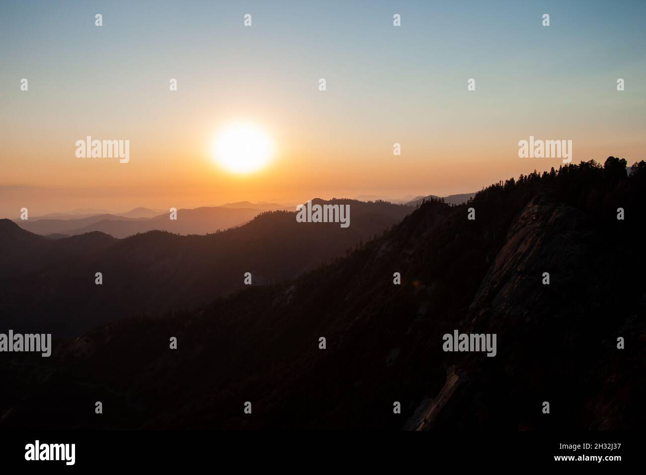 Wunderschöner Sonnenuntergang über den Bergen, die von Wald überwuchert sind, riesige orangefarbene Sonne geht unter, blauer Himmel | szenischer Sonnenuntergang am Moro Rock im Sequoia National Park USA Stockfoto