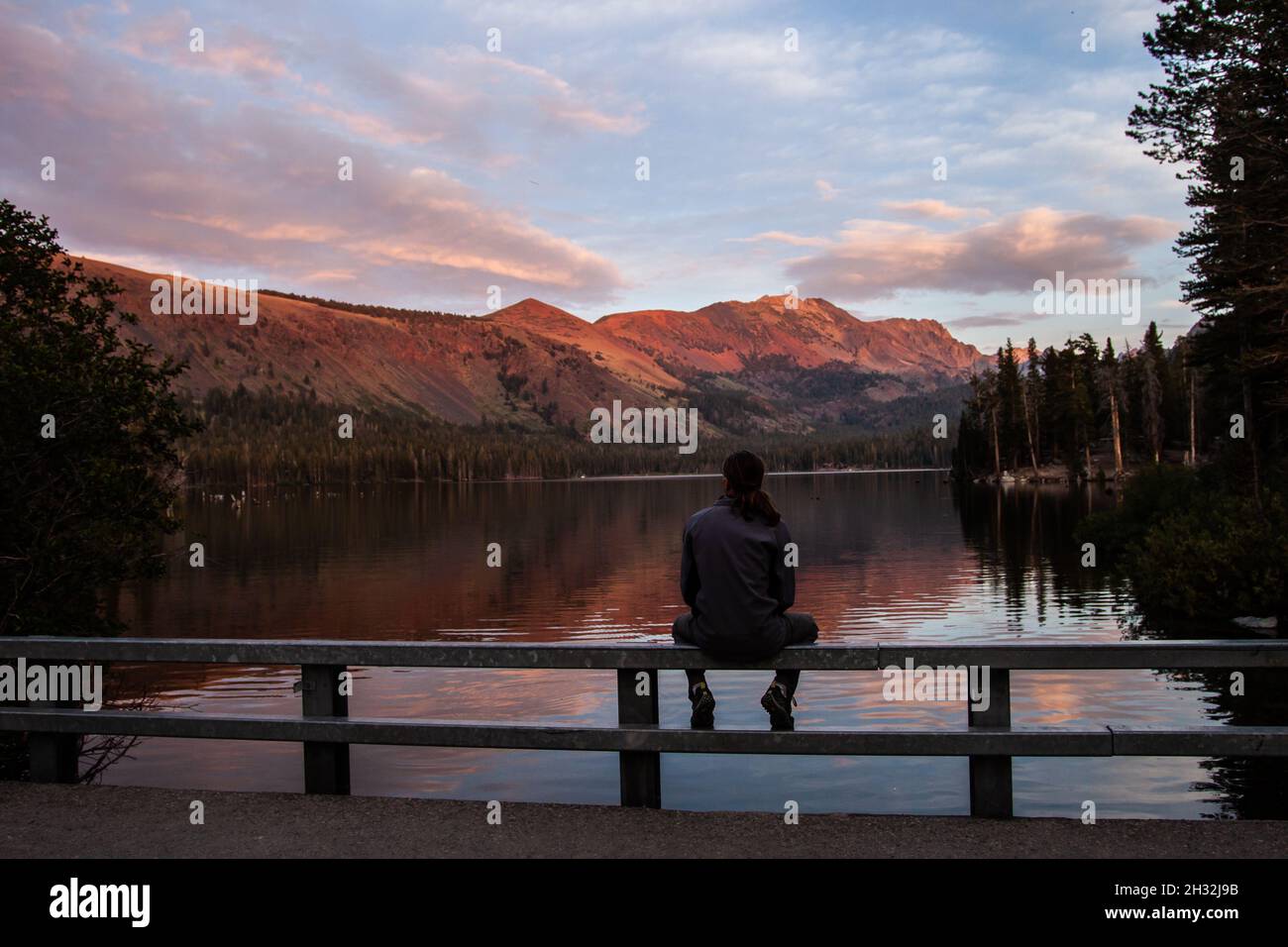 Mann sitzt auf dem Geländer und schaut auf Lake Mary (Mammoth Lakes) bei Sonnenuntergang, Kalifornien, Berge spiegeln sich im Wasser, See umgeben von Bäumen, Blick auf Serene Stockfoto