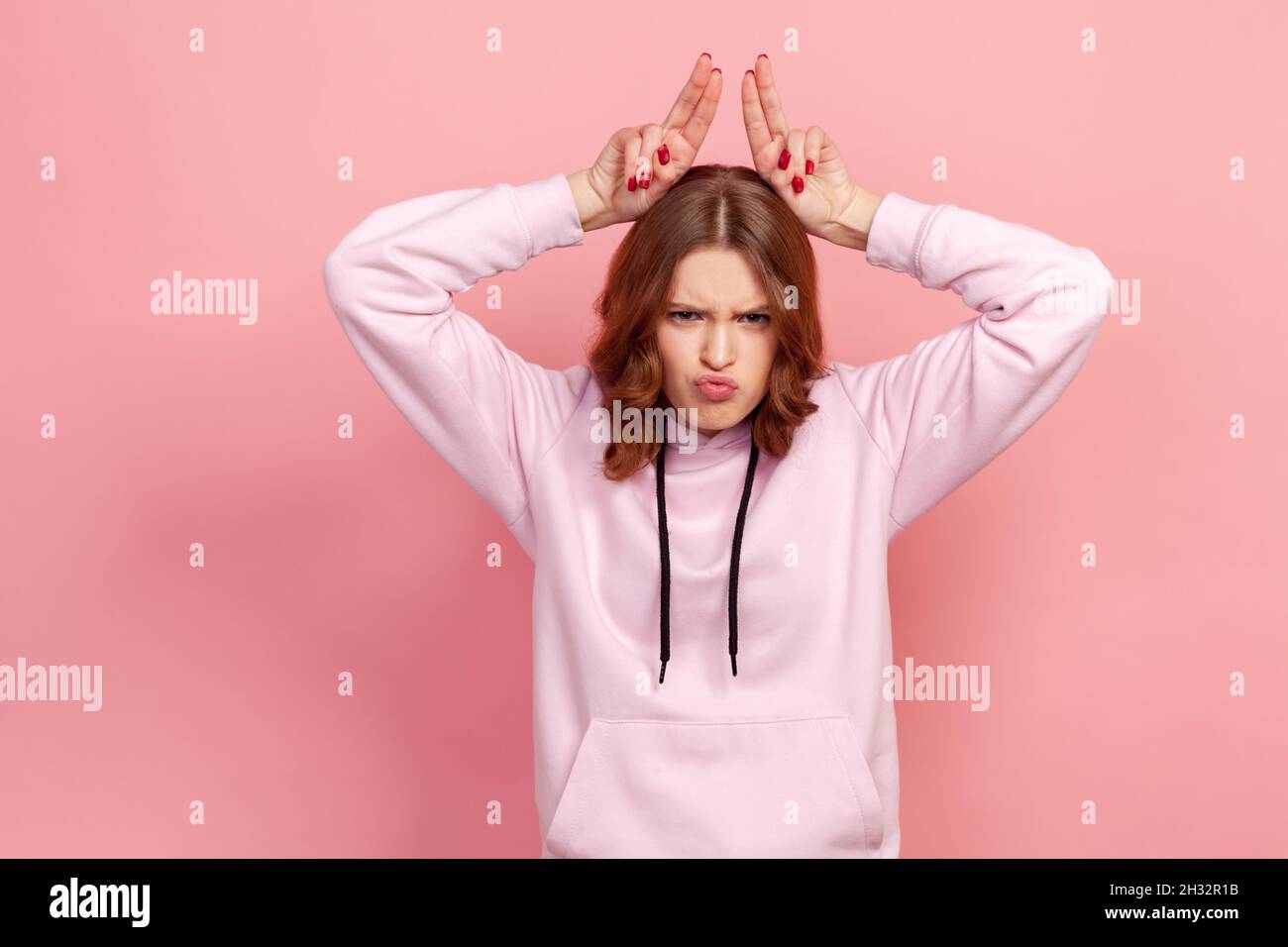 Portrait eines aggressiven Teenagers im Kapuzenpullover mit Stierhörnern, Geweihkopfgesten, das zu Angriffen, Konflikten oder Verteidigungskonzepten droht. Innenaufnahme des Studios isoliert auf rosa Hintergrund Stockfoto