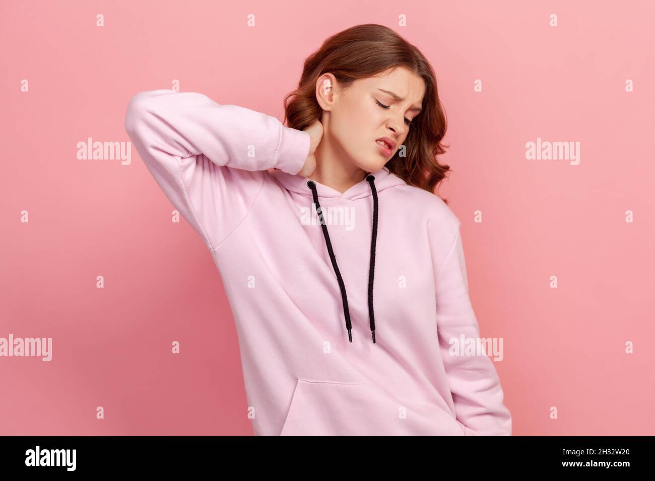 Porträt von verärgert Brünette Teenager-Mädchen im Hoodie berühren Hals Gefühl Schmerzen und Taubheit, besorgt über Muskelverspannungen, Osteochondrose. Innenaufnahme des Studios isoliert auf rosa Hintergrund Stockfoto
