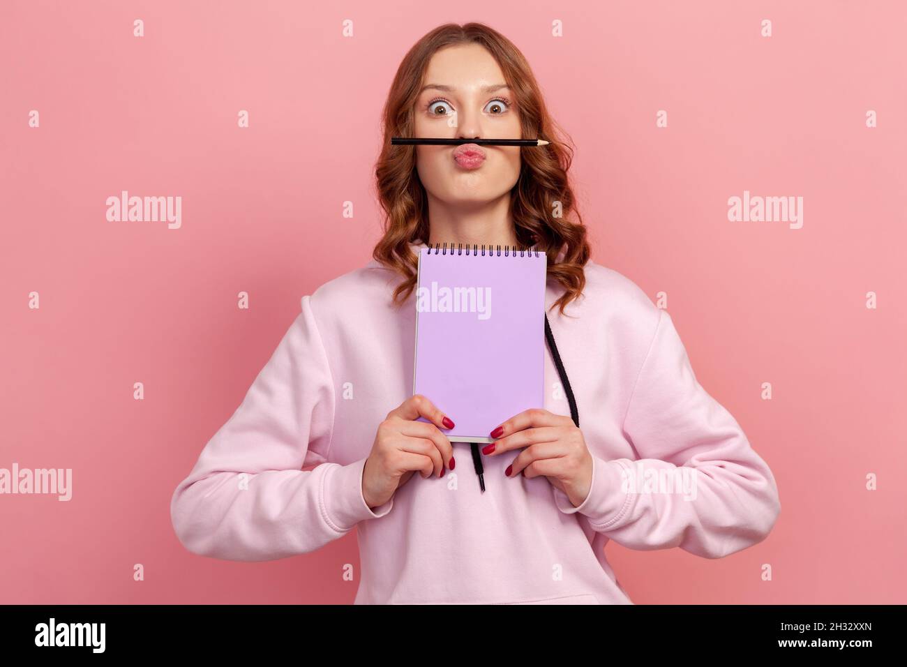 Porträt eines lustigen lockigen Teenagers im Hoodie mit Bleistift unter der Oberlippe, das Notizbuch zeigt, mit großen Augen auf die Kamera schaut und herumtollt. Innenaufnahme des Studios isoliert auf rosa Hintergrund Stockfoto