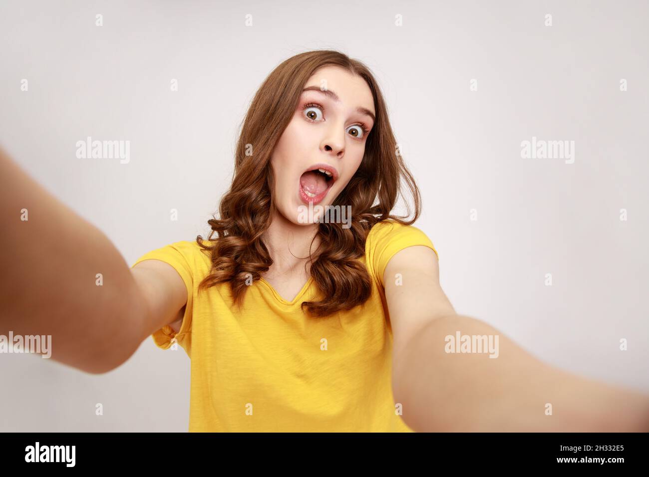 Porträt eines aufgeregt überraschten Teenagers, das ein gelbes T-Shirt trägt, das Selfie macht, die Kamera mit offenem POV anschaut, aus der Perspektive des Fotos. Innenaufnahme des Studios isoliert auf grauem Hintergrund. Stockfoto