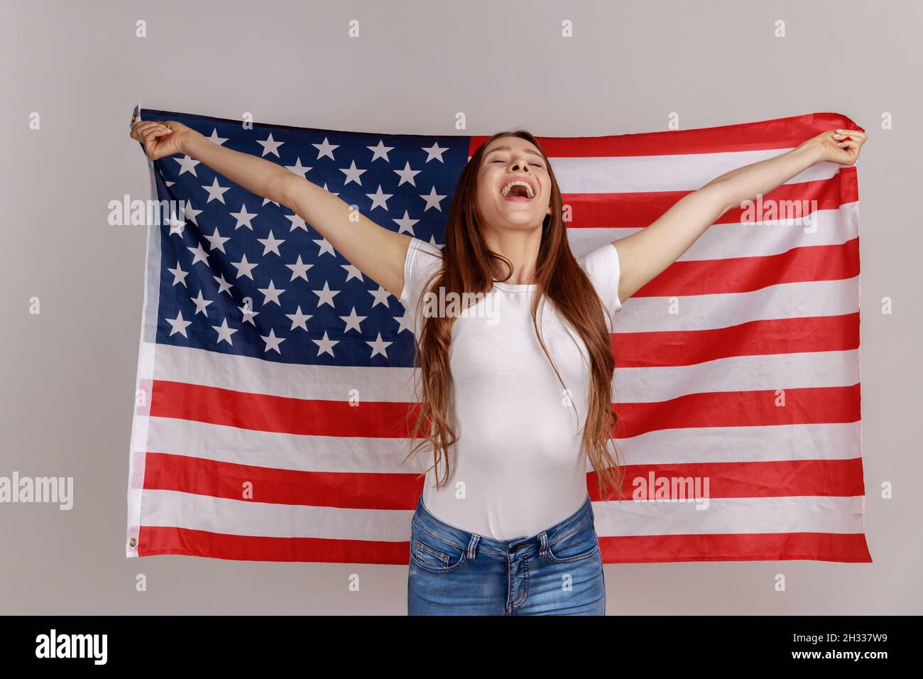 Porträt einer attraktiven, schönen jungen Frau mit dunklem Haar, die die USA-Flagge über den Schultern hält und sich glücklich freut, das weiße T-Shirt trägt. Innenaufnahme des Studios isoliert auf grauem Hintergrund. Stockfoto