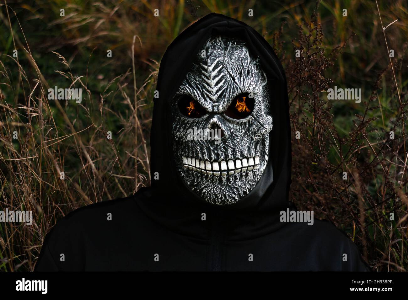 Nahaufnahme des grimmigen Reaper. Mann in Totenmaske mit Feuerflamme in den Augen auf dunklem Natur trockenem Gras Hintergrund. Faschingskostüm, gruselige Zähne. Hallo Stockfoto