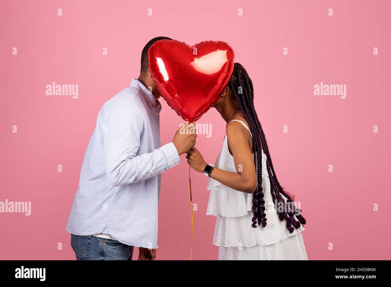 Afroamerikanisches Paar in Liebe küsst, hält und versteckt sich hinter einem roten herzförmigen Ballon, bedeckt ihre Gesichter für Privatsphäre, isoliert auf rosa Hintergrund Stockfoto