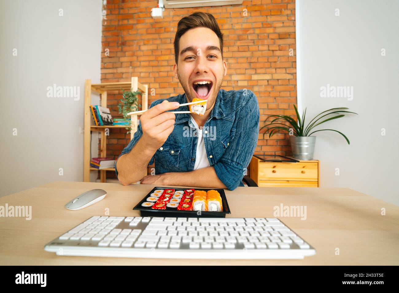 Vorderansicht eines fröhlichen, hübschen jungen Mannes, der Sushi isst und Informationen vom Monitor liest und die Kamera anschaut. Stockfoto