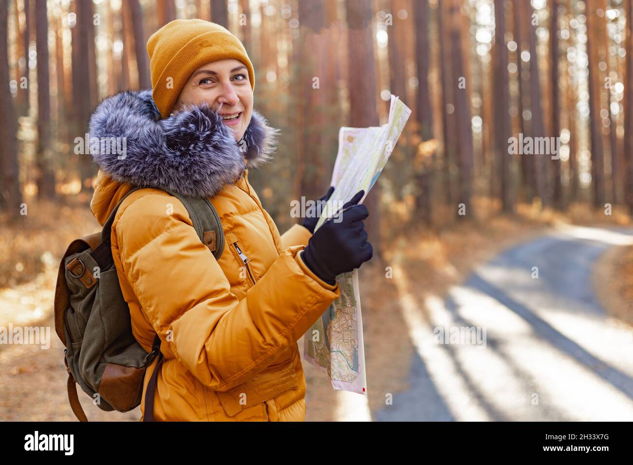 Junge lächelnde Frau in einer warmen orangefarbenen Jacke. Attraktive kaukasische Ethnie Frau spaziert im Park mit einem Rucksack. Sie schaut sich die Karte an. Wandern in t Stockfoto